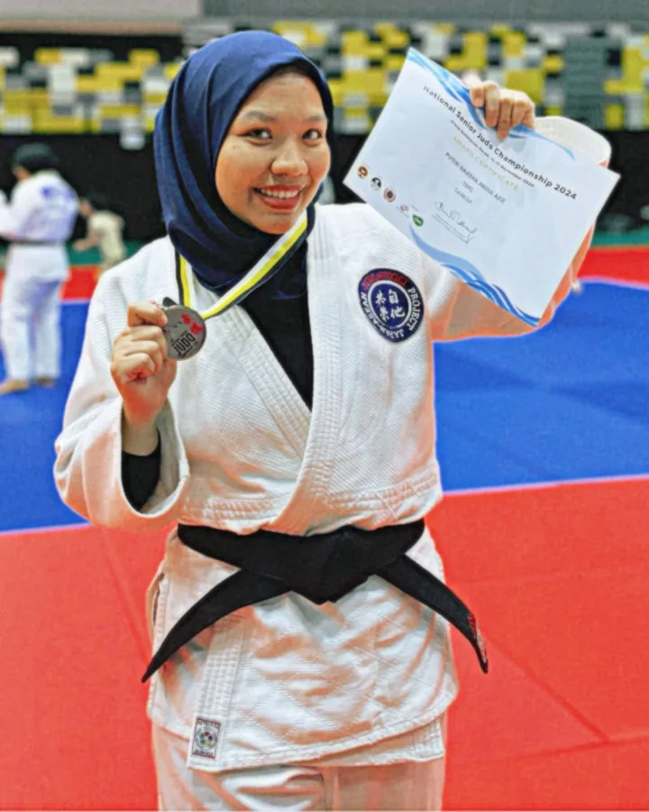 Coach Puteri in a white judo gi and black belt, holding a medal and a National Senior Judo Championship 2024 certificate